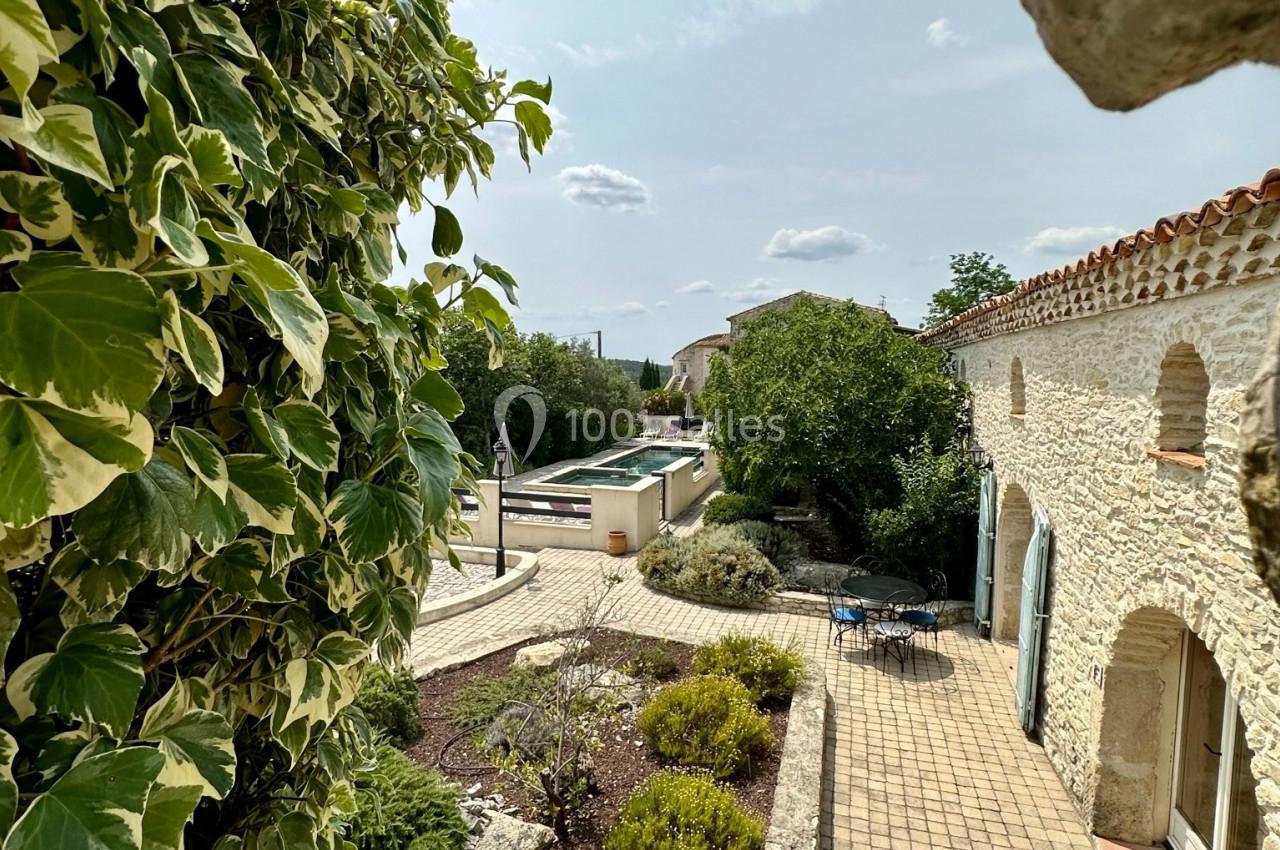 Vue d'une cour en pierre avec terrasse, végétation, piscine et ciel dégagé, encadrée par un mur en premier plan.