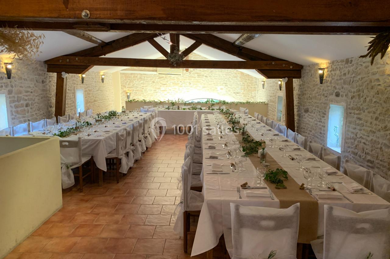 Salle de réception avec deux longues tables dressées, décorées de nappes blanches et de feuillages, dans un cadre en pierre.