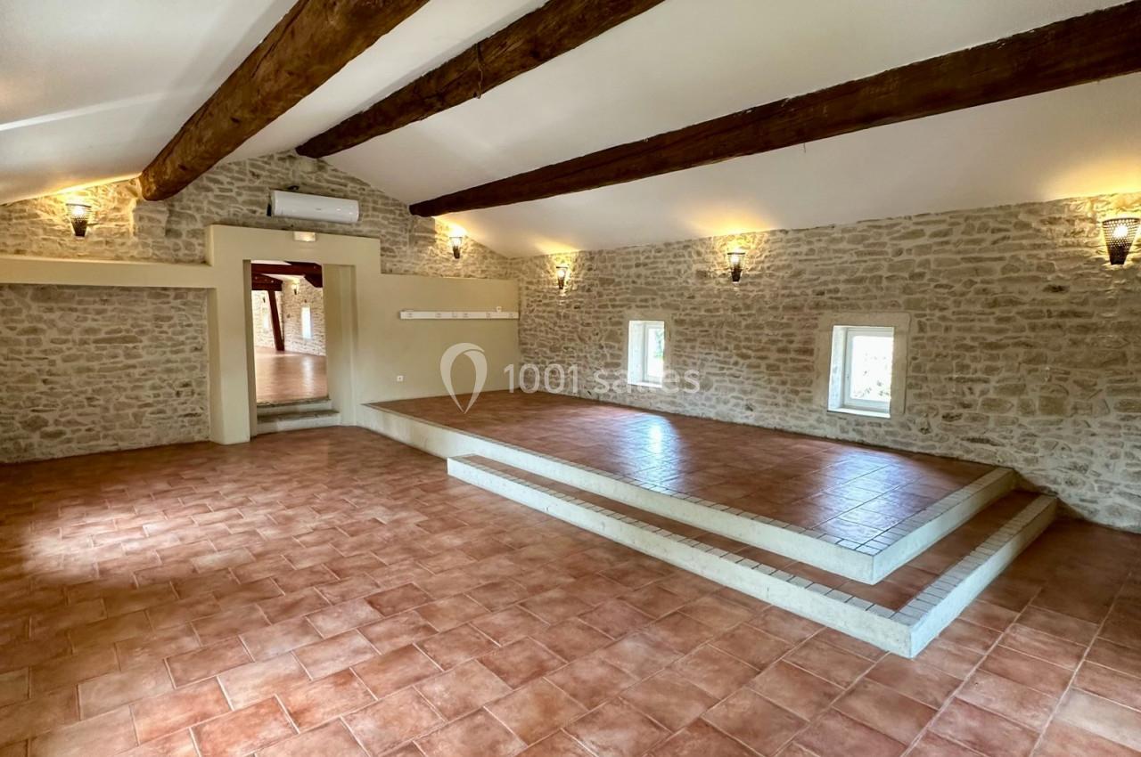 Salle lumineuse avec murs en pierre, poutres apparentes, sol en carrelage et estrade surélevée.