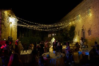 Dîner en plein air de nuit avec des guirlandes lumineuses, des invités assis autour de tables dans une cour en pierre.