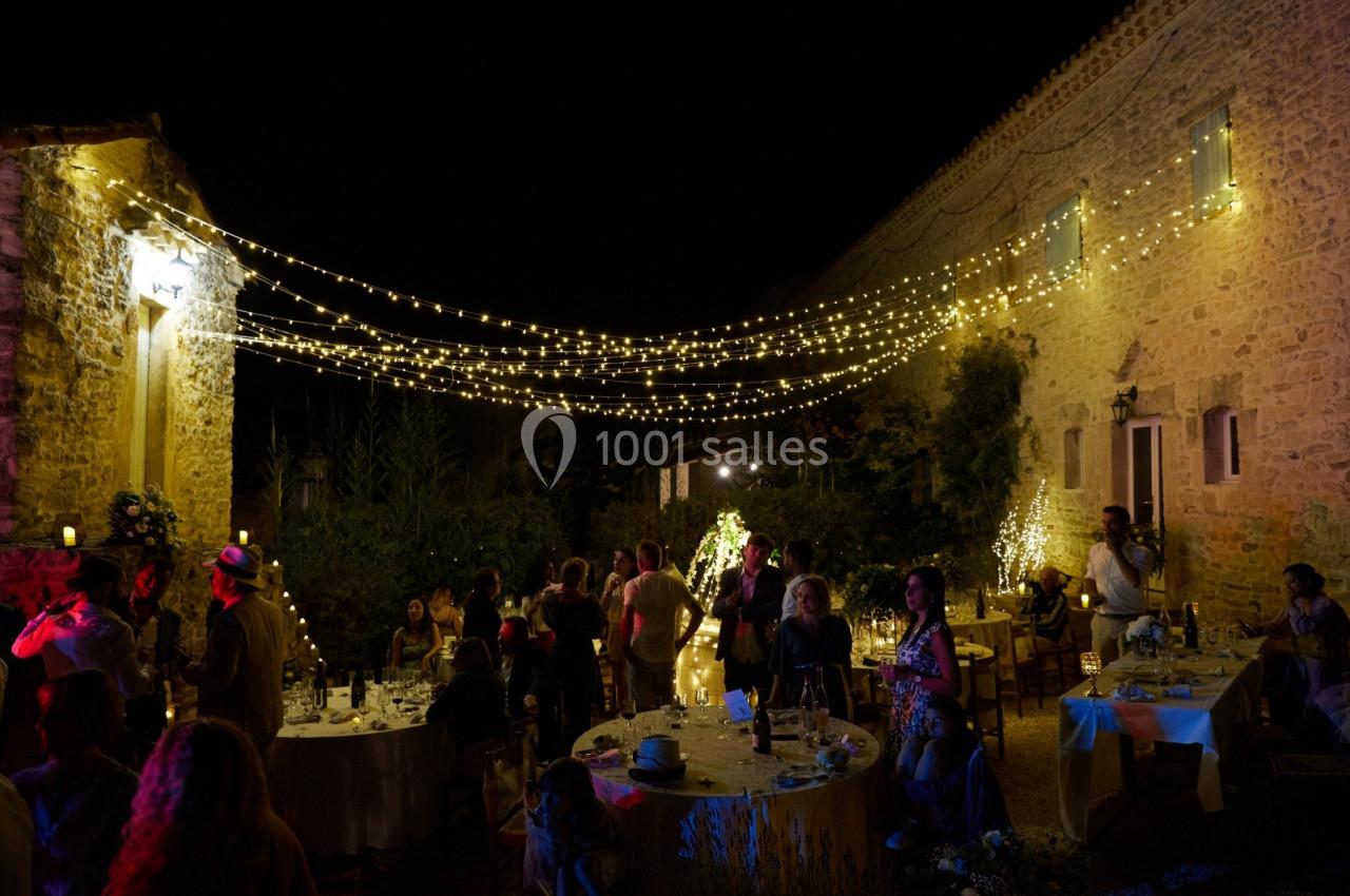 Cour extérieure éclairée par des guirlandes lumineuses, avec des invités assis et debout lors d'une réception nocturne.