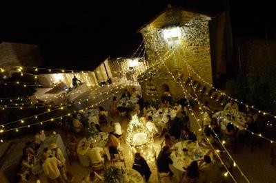 Dîner en plein air de nuit avec des guirlandes lumineuses, des invités assis autour de tables dans une cour en pierre.