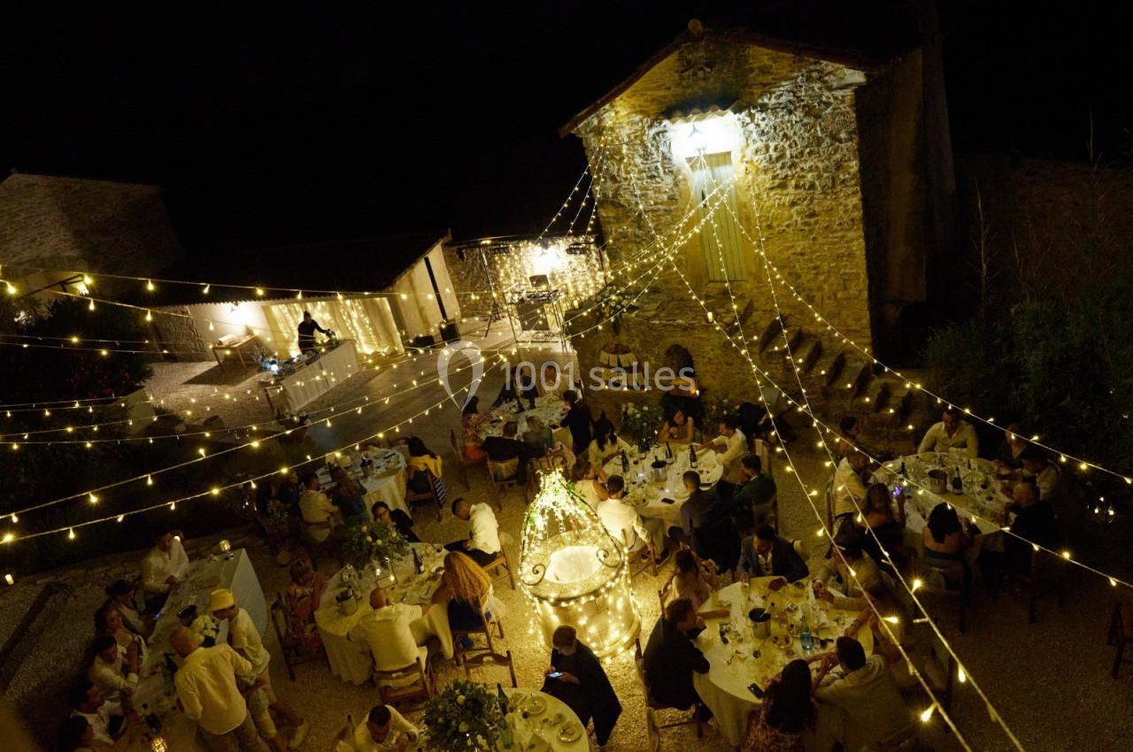 Dîner en plein air de nuit avec des guirlandes lumineuses, des invités assis autour de tables dans une cour en pierre.
