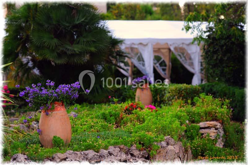 Jardin fleuri avec des jarres en terre cuite, des plantes colorées et une tente blanche en arrière-plan.