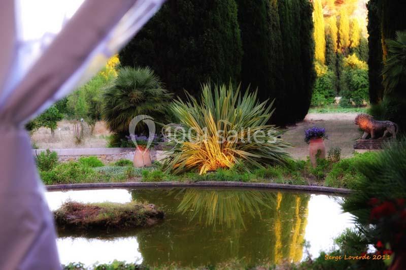 Vue d'un jardin méditerranéen avec un bassin, des plantes luxuriantes et des arbres en arrière-plan.