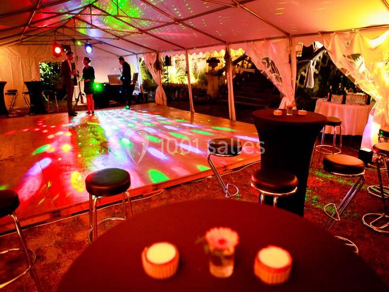 Salle de réception sous une tente éclairée par des lumières colorées, avec piste de danse et tables hautes.