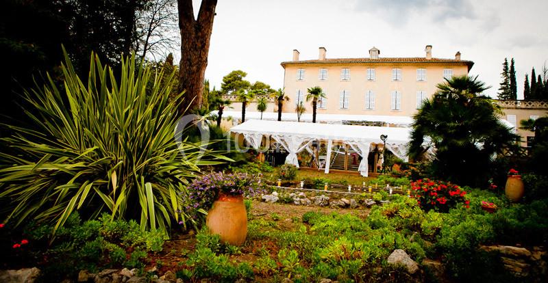Jardin fleuri avec plantes méditerranéennes, vue sur une grande bâtisse en arrière-plan sous un ciel nuageux.