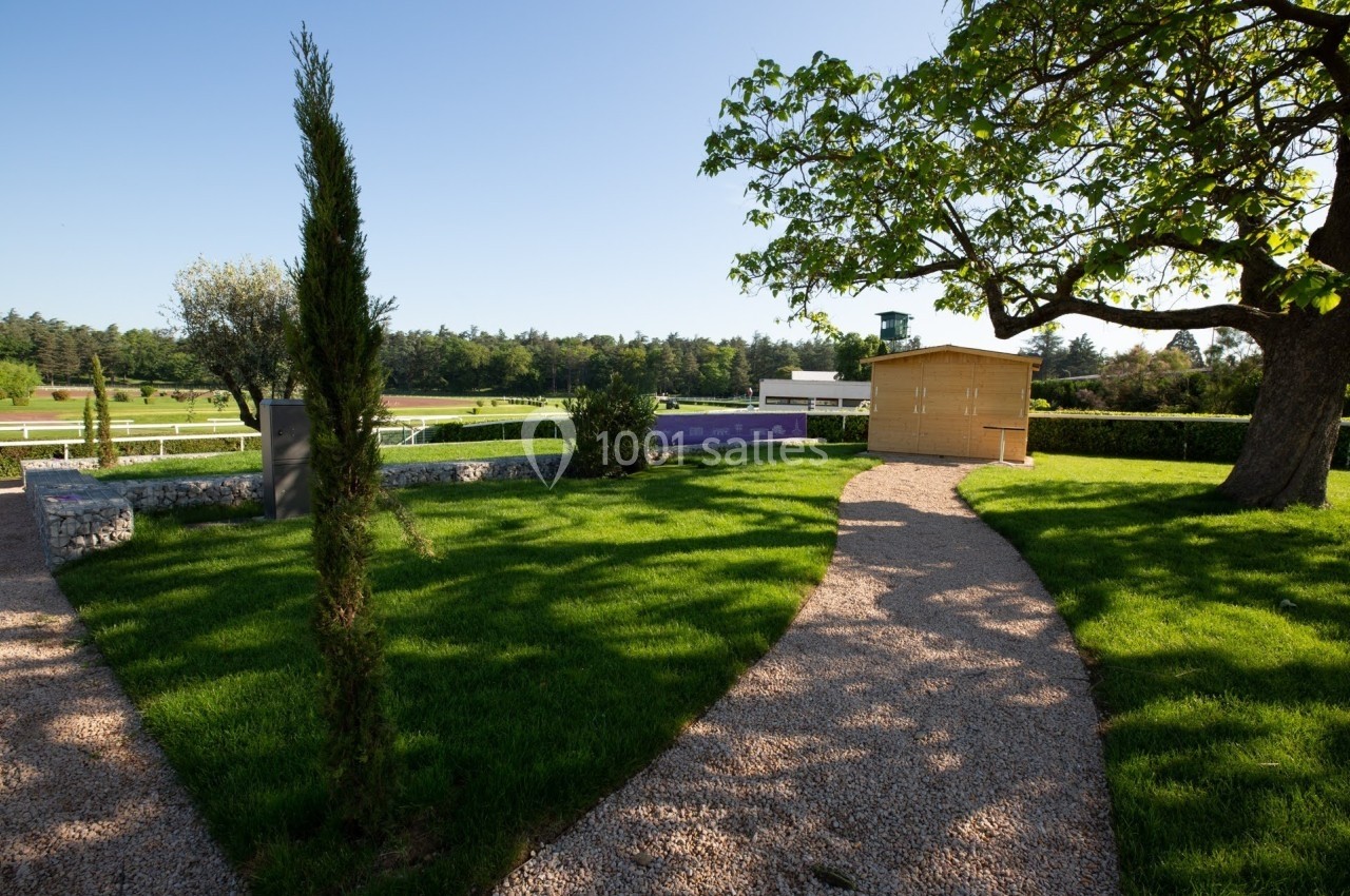 Allée gravillonnée serpentant à travers un jardin verdoyant avec un arbre, un cabanon en bois et une clôture au fond.