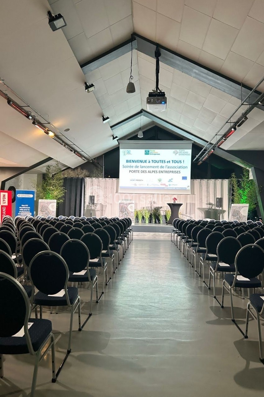 Salle de conférence avec rangées de chaises vides, scène équipée d'un écran de projection et décor végétal en arrière-plan.