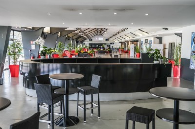 Salle avec grandes baies vitrées donnant sur une piste extérieure, aménagée avec tables, chaises et fauteuils rouges.