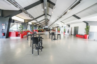 Salle avec grandes baies vitrées donnant sur une piste extérieure, aménagée avec tables, chaises et fauteuils rouges.