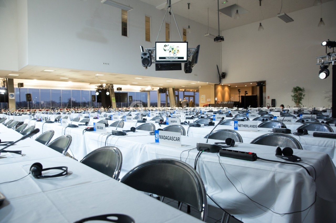Salle de conférence avec des rangées de tables et de chaises équipées de casques audio, étiquettes de pays visibles.