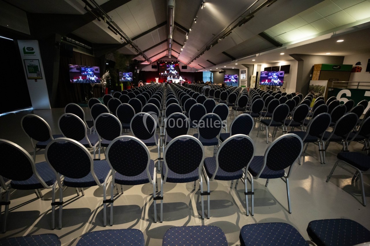 Salle de conférence avec des rangées de chaises vides, scène éclairée et écrans affichant des présentations.