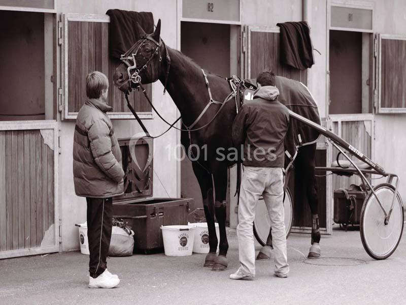 Deux personnes préparent un cheval attelé devant des boxes dans une écurie.