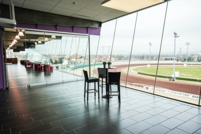 Salle avec grandes baies vitrées donnant sur une piste extérieure, aménagée avec tables, chaises et fauteuils rouges.