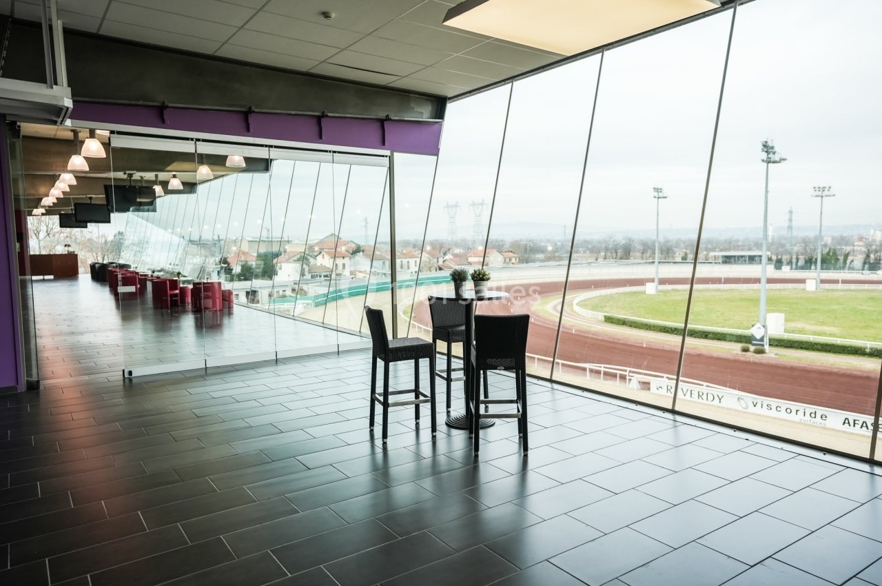 Vue intérieure d'une salle avec grandes baies vitrées donnant sur une piste d'hippodrome et des gradins extérieurs.