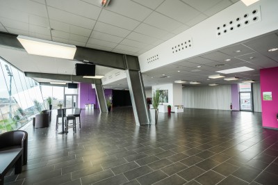 Salle avec grandes baies vitrées donnant sur une piste extérieure, aménagée avec tables, chaises et fauteuils rouges.