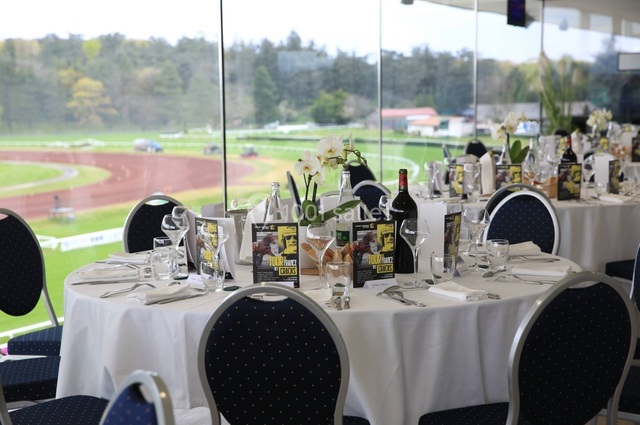 Tables élégamment dressées avec nappes blanches, verres et couverts, donnant sur une piste de course visible à travers de…