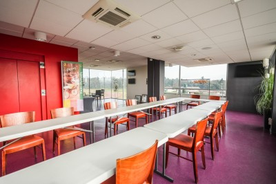Salle avec grandes baies vitrées donnant sur une piste extérieure, aménagée avec tables, chaises et fauteuils rouges.