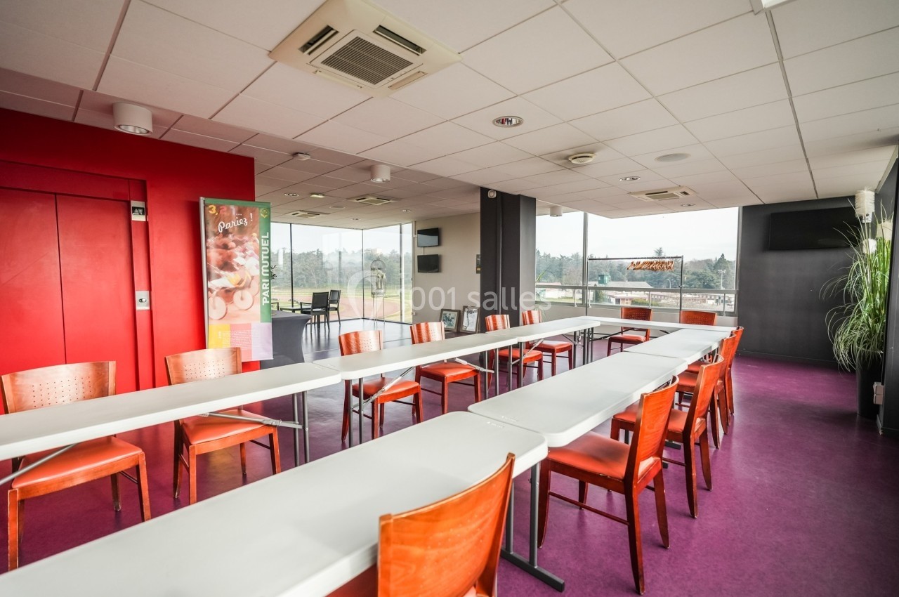 Salle lumineuse avec de grandes baies vitrées, tables blanches alignées et chaises en bois sur un sol violet.