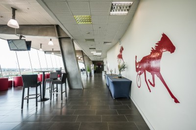 Salle avec grandes baies vitrées donnant sur une piste extérieure, aménagée avec tables, chaises et fauteuils rouges.