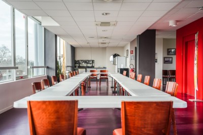 Salle avec grandes baies vitrées donnant sur une piste extérieure, aménagée avec tables, chaises et fauteuils rouges.
