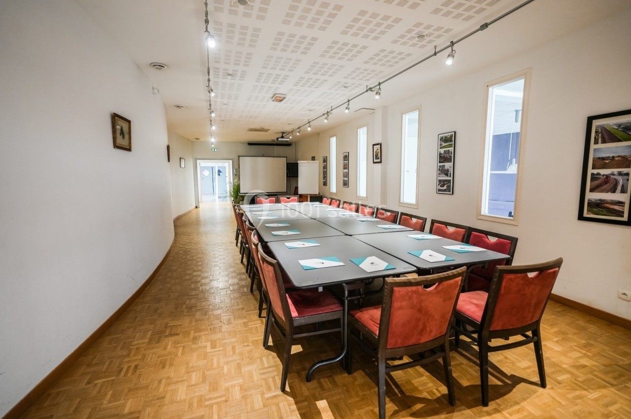 Salle de réunion lumineuse avec une grande table rectangulaire entourée de chaises rouges et un projecteur au fond.