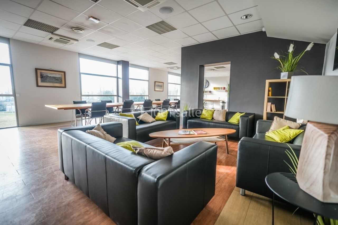 Salle lumineuse avec canapés noirs, coussins colorés, table basse en bois, étagère et grandes fenêtres.