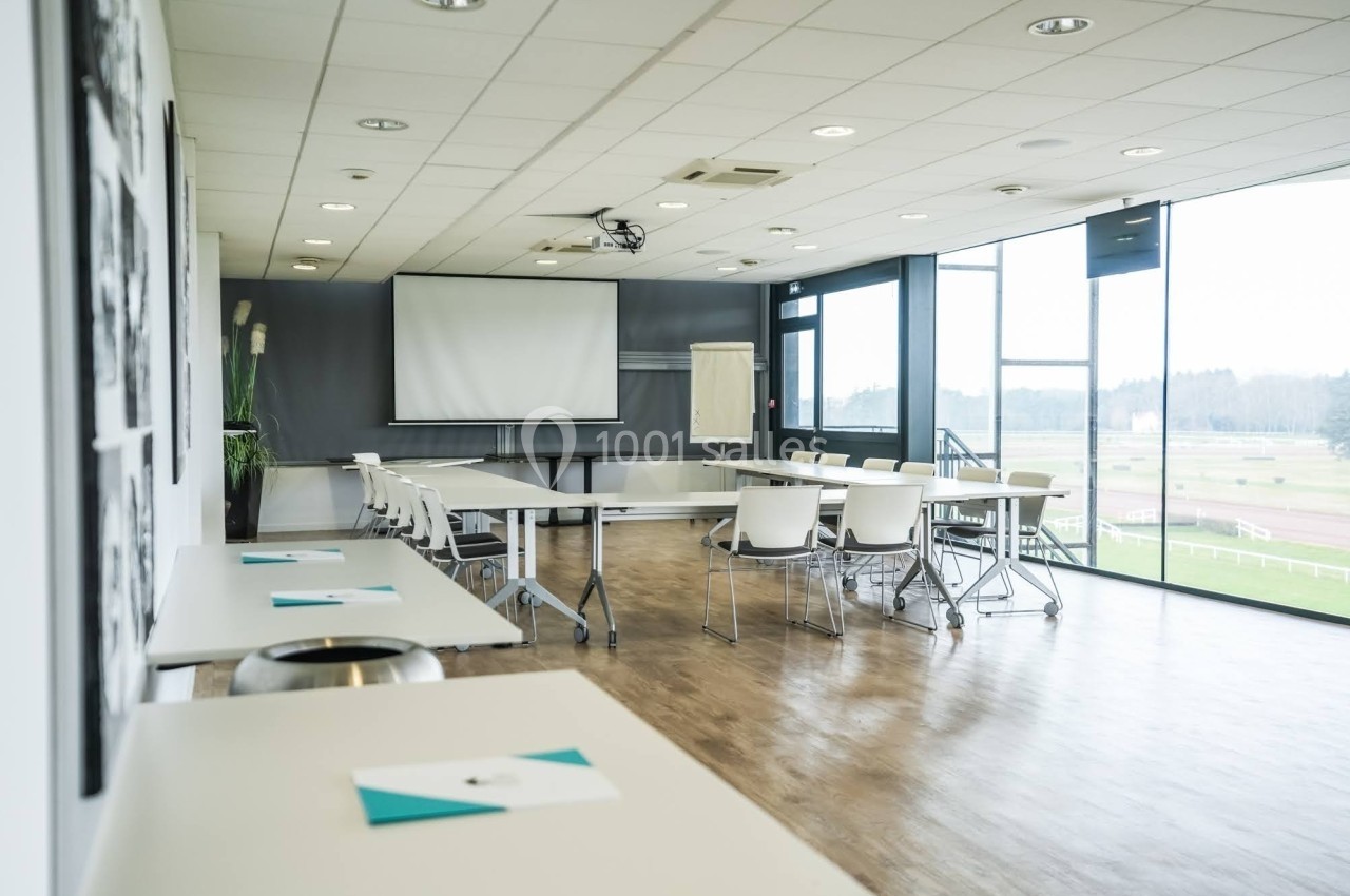 Salle de réunion lumineuse avec tables, chaises blanches, écran de projection et vue sur un espace extérieur verdoyant.