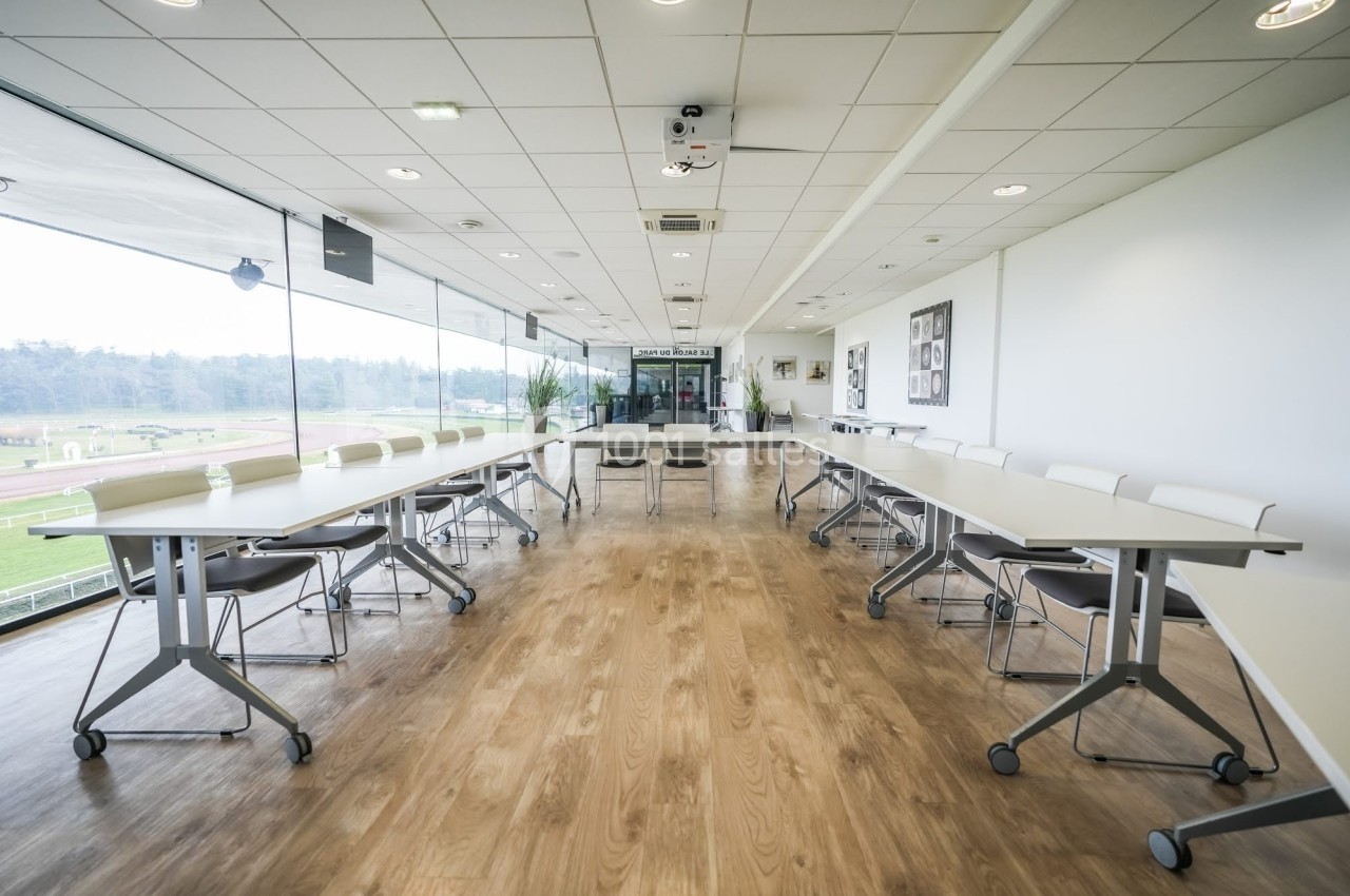 Salle de réunion lumineuse avec grandes baies vitrées, tables en U et sol en bois clair.