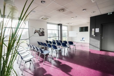 Salle avec grandes baies vitrées donnant sur une piste extérieure, aménagée avec tables, chaises et fauteuils rouges.
