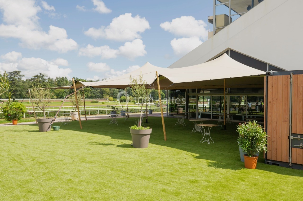 Terrasse extérieure avec tables et chaises sous une tente, entourée de plantes, sur une pelouse verte par temps ensoleillé.