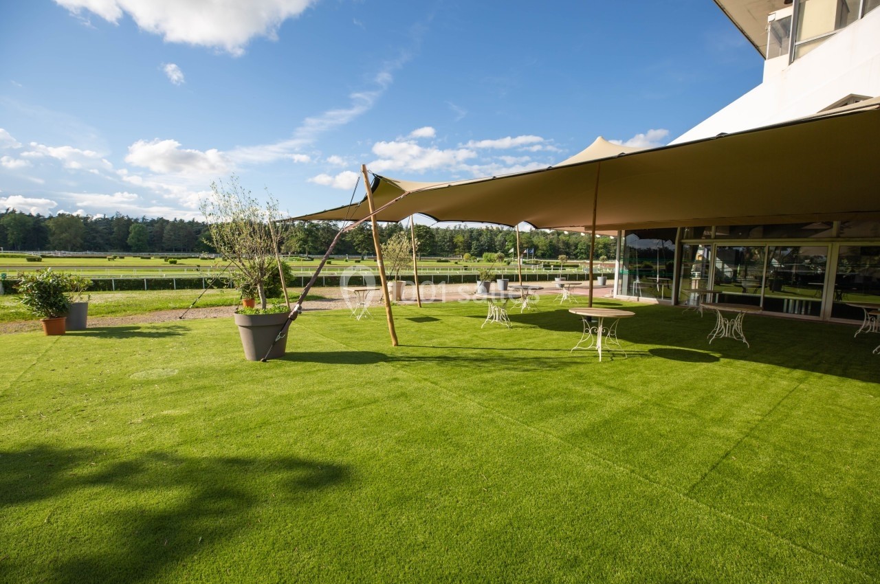 Espace extérieur avec pelouse, tables et chaises sous des toiles d'ombrage, entouré de verdure et d'un ciel dégagé.