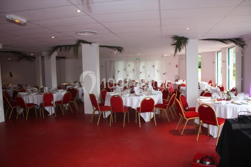 Salle de réception avec tables rondes dressées, nappes blanches, chaises rouges et décorations végétales au plafond.