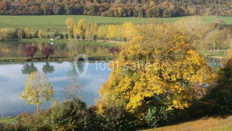 Paysage automnal avec un lac reflétant des arbres aux feuilles jaunes, rouges et vertes, entouré de champs et collines.
