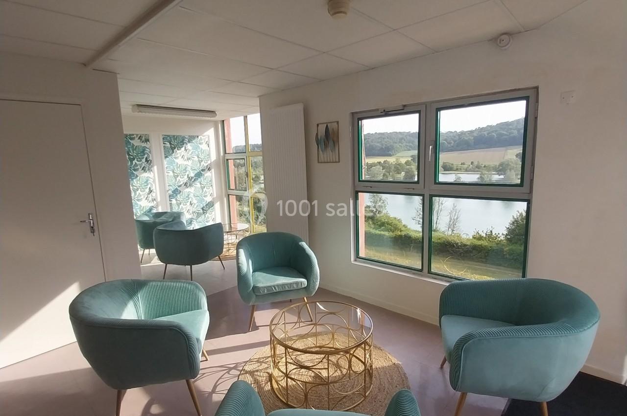 Espace lumineux avec fauteuils verts, table basse dorée et vue sur un paysage extérieur à travers de grandes fenêtres.