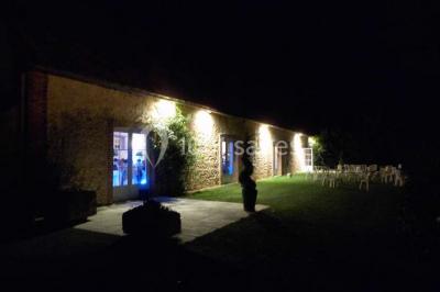 Façade d'un bâtiment en pierre éclairée la nuit, avec des portes vitrées et des chaises disposées sur une pelouse.