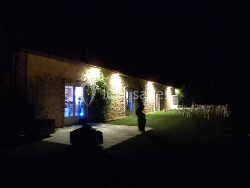 Façade d'un bâtiment en pierre éclairée la nuit, avec des portes vitrées et des chaises disposées sur une pelouse.