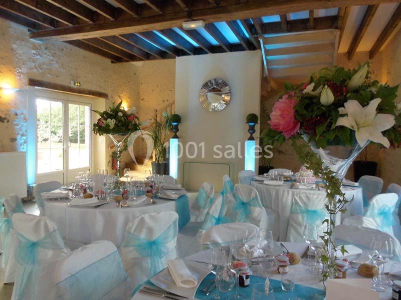 Salle de réception décorée avec des tables rondes, nappes blanches, chaises ornées de nœuds bleus et compositions florales.