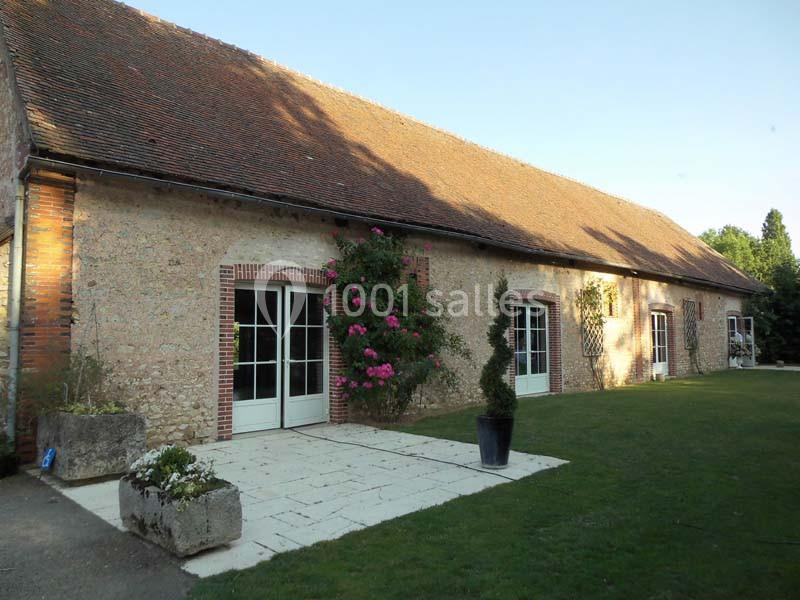 Façade d'une longère en pierre avec toit en tuiles, rosiers grimpants et terrasse pavée donnant sur une pelouse.