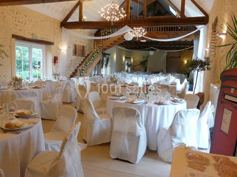 Salle de réception décorée avec des tables rondes dressées, chaises blanches et poutres apparentes en bois.
