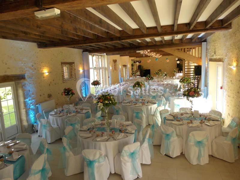 Salle de réception décorée pour un mariage, avec tables rondes, nappes blanches et chaises ornées de nœuds bleus.