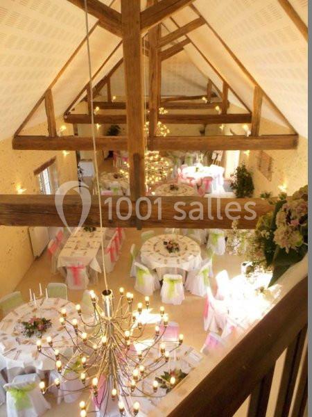 Salle de réception lumineuse avec poutres apparentes, tables rondes décorées et grand lustre central.