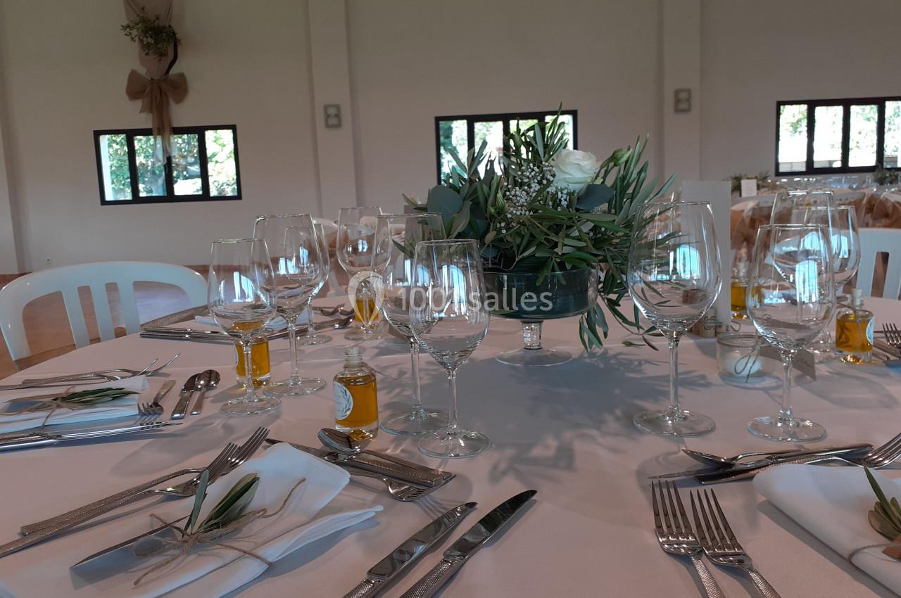 Table dressée pour un événement avec nappes blanches, couverts, verres, décorations végétales et petites bouteilles d'huile.
