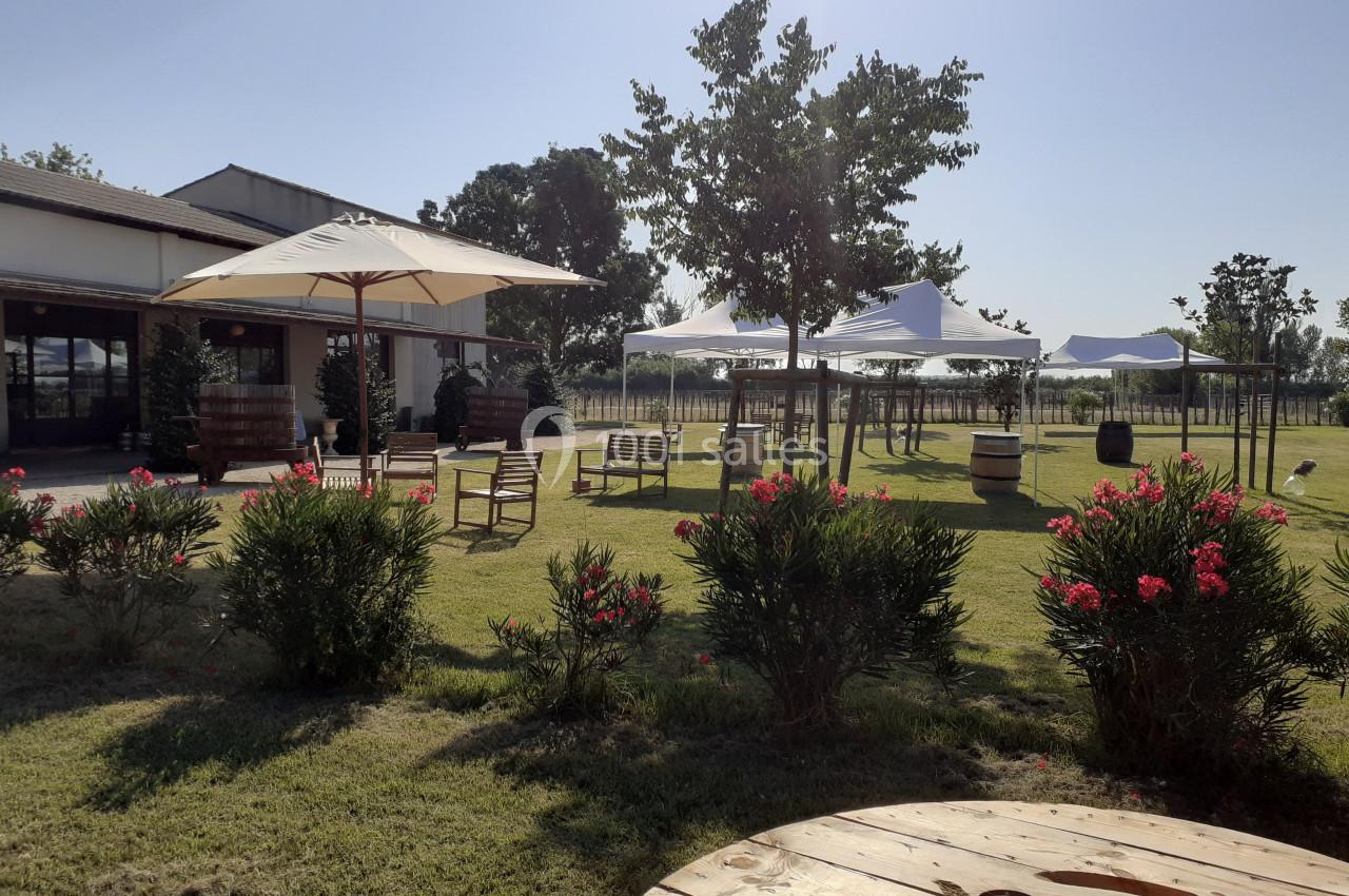 Espace extérieur avec pelouse, parasols, tables, chaises et arbustes fleuris, sous un ciel dégagé.