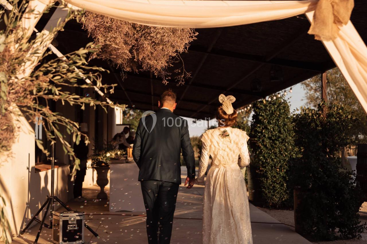 Un couple en tenue de mariage marche sous une arche décorée, éclairée par une lumière de fin de journée.