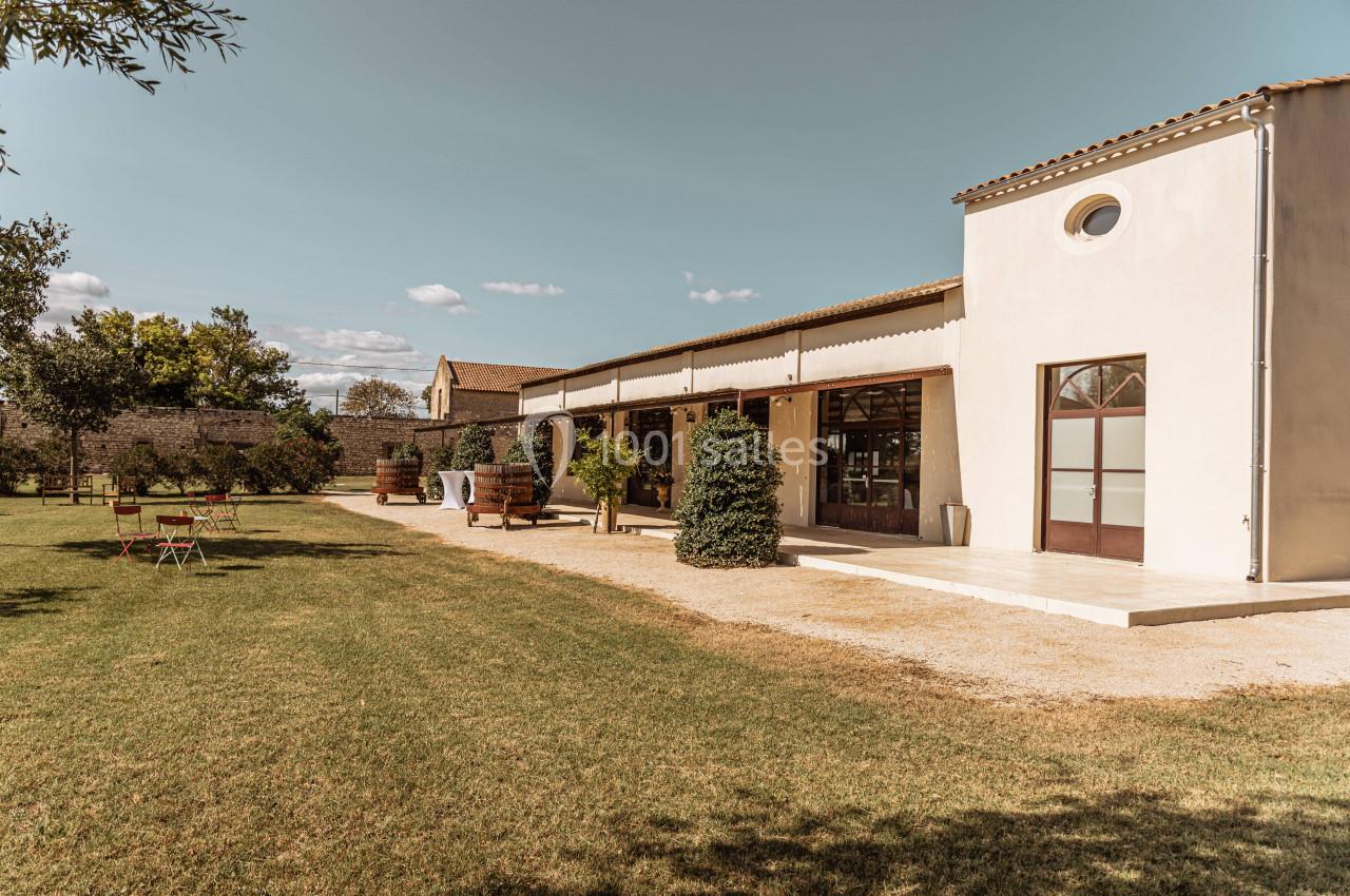 Bâtiment clair avec terrasse, entouré de pelouse, arbres et mobilier de jardin sous un ciel dégagé.