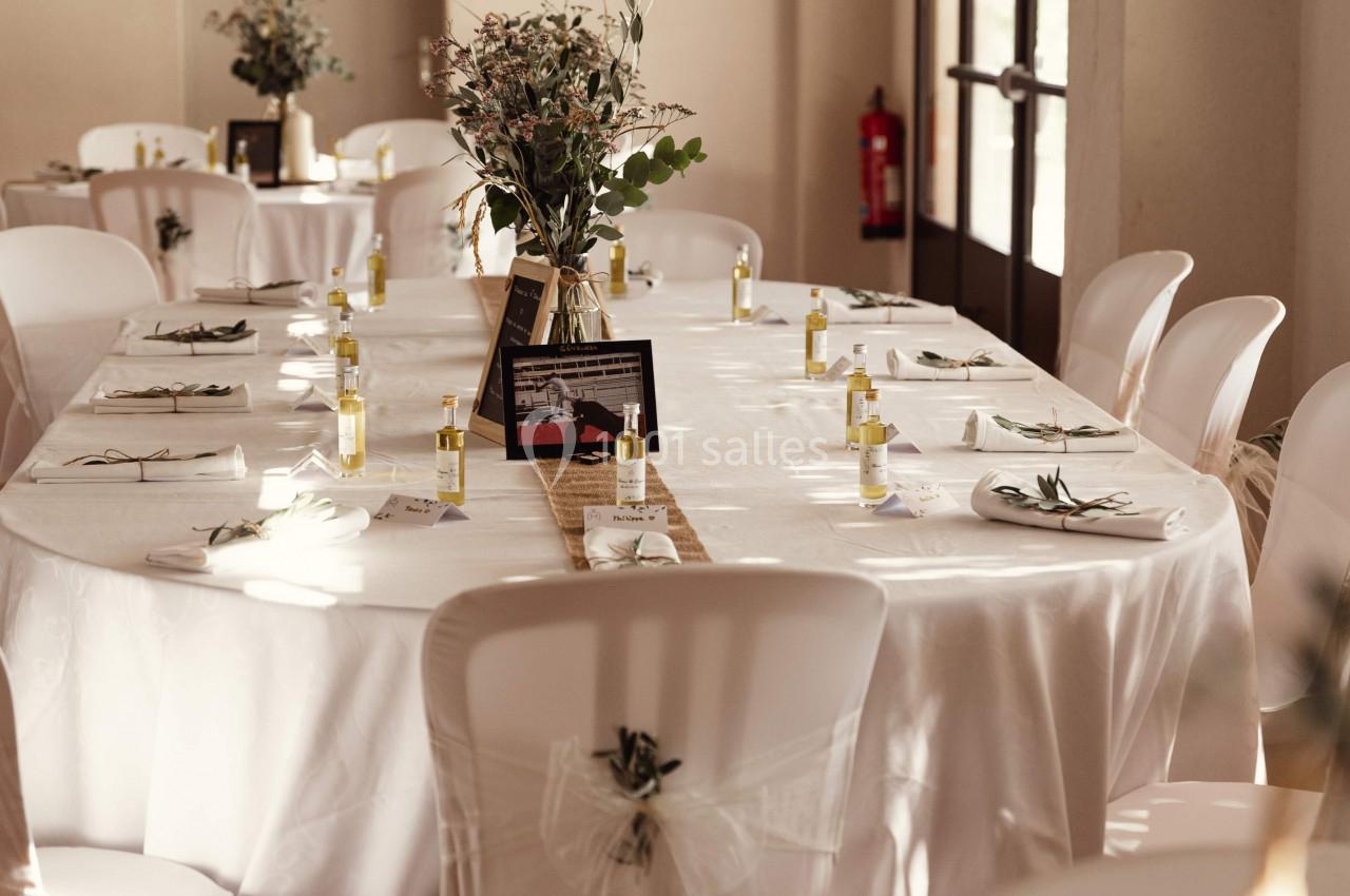 Salle de réception avec tables ovales dressées, nappes blanches, chaises décorées et centres de table floraux.
