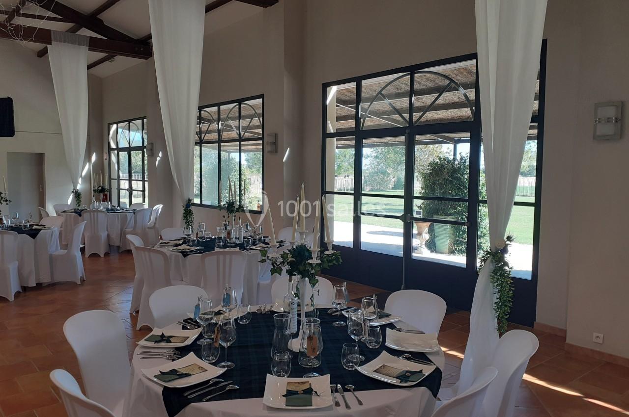 Salle de réception lumineuse avec tables dressées, nappes sombres, chaises blanches et grandes baies vitrées donnant sur un…