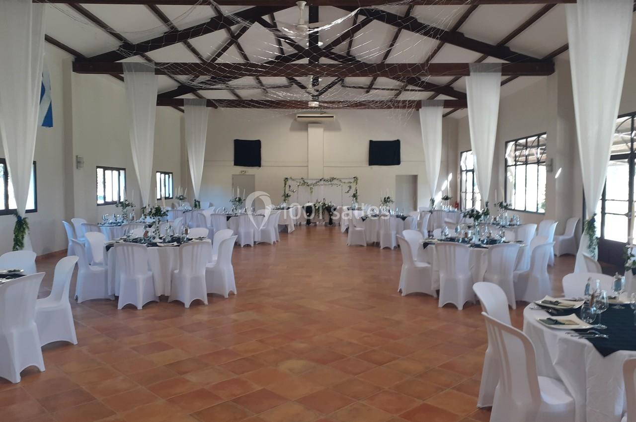 Salle de réception décorée avec des tables rondes dressées, nappes blanches et chaises assorties, sous un plafond en poutres…